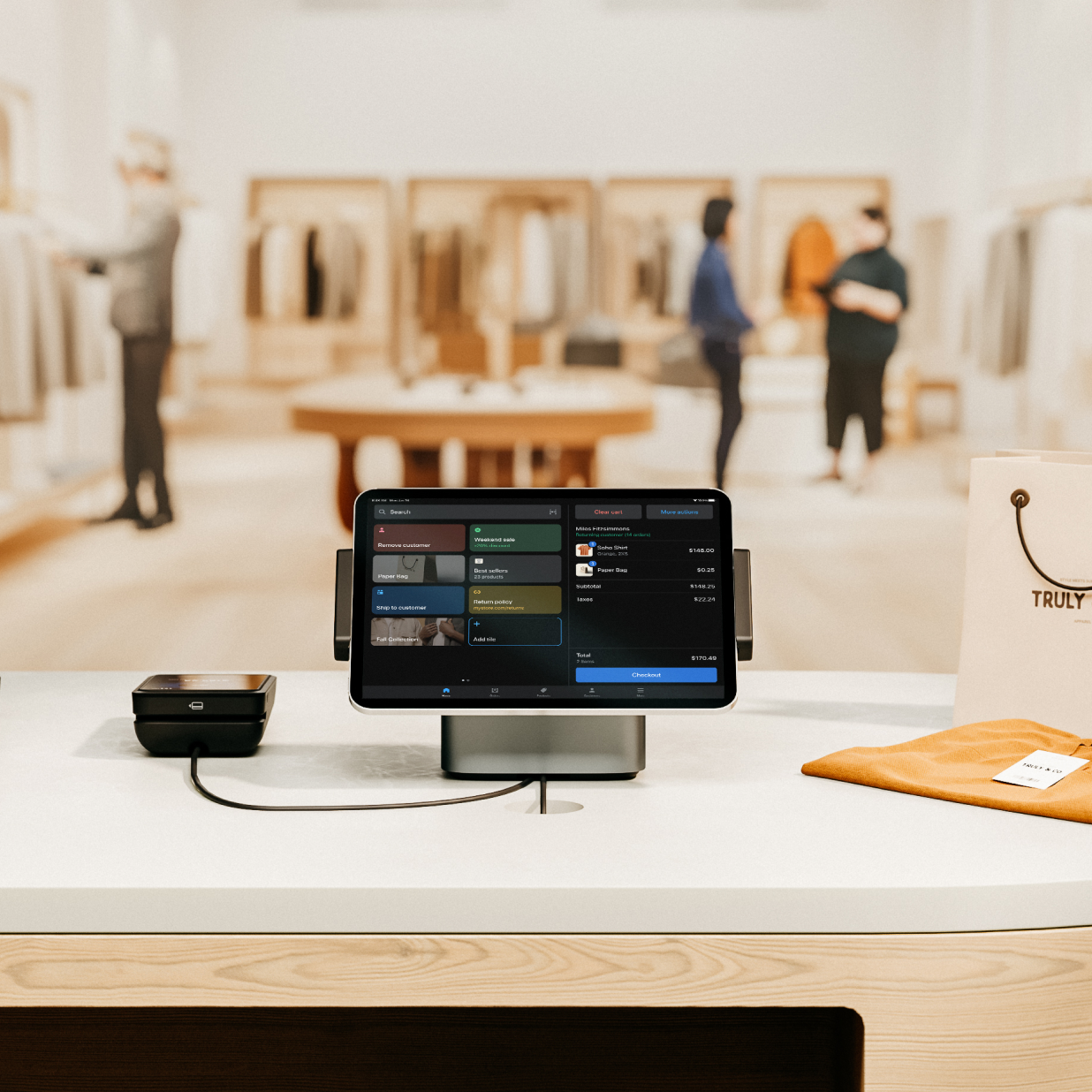 A view from behind the counter of a large, apparel store called Truly and Co, in a beautiful and brightly lit mall. On the counter there are two Shopify Point of Sale setups that show the POS app on a tablet, which is connected directly to POS Terminal. In the store are people shopping and a sales associate helping a customer checkout on the floor.